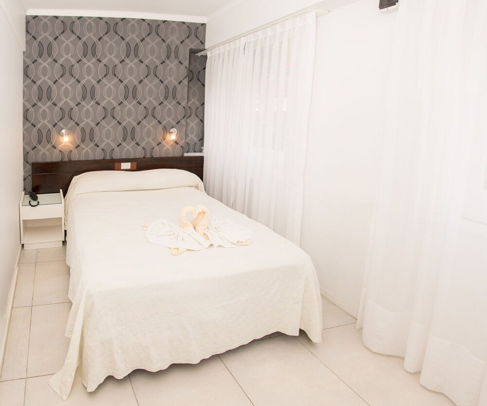 Cama prolija con ropa blanca, mesa de luz con lámpara y empapelado