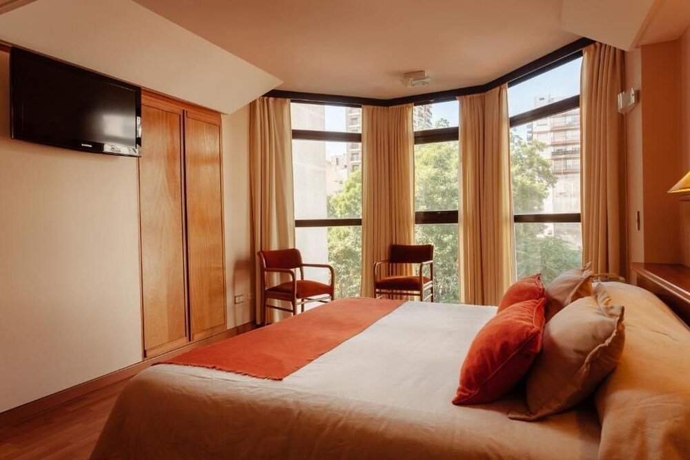 Habitación con cama grande, dos sillas, TV y ventanales con vista a edificios